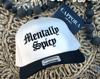 Puede incluir: Una gorra de béisbol blanca con visera negra y el texto Mentally Spicy bordado en letras góticas negras en la parte delantera. Una pequeña pegatina rectangular está adherida a la visera y se ve una etiqueta de marca azul oscuro con el texto Cappora Apparel.