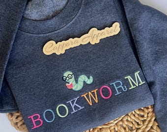 Puede incluir: Una sudadera de cuello redondo en gris jaspeado oscuro con un gusano verde claro bordado que lleva gafas negras y lee un pequeño libro rosa. La palabra BOOKWORM está bordada en letras multicolores debajo del gusano, con una etiqueta de madera.