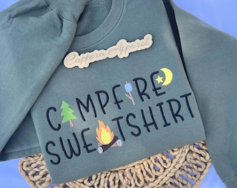 Puede incluir: Una sudadera de cuello redondo color verde oliva con texto bordado en negro que dice CAMPFIRE SWEATSHIRT. El diseño incluye un pino verde, un malvavisco azul en un palo, una luna creciente amarilla con una estrella y una fogata con llamas naranjas.
