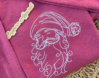 Puede incluir: Una sudadera roja jaspeada que presenta un diseño bordado en hilo blanco de una cara de Papá Noel con gorro y barba. Una etiqueta de madera con el texto Cappora Apparel está colocada sobre la tela junto al gráfico bordado.