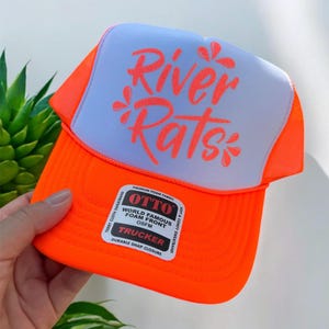 Peut inclure: Une casquette de camionneur orange néon vif avec un panneau avant en mousse blanche portant le texte River Rats imprimé dans une police cursive orange vif. La casquette comprend un dos en maille, une visière incurvée et une étiquette rectangulaire.