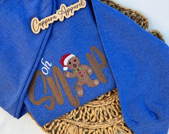 Puede incluir: Una sudadera azul de manga larga con un hombre de jengibre bordado que lleva un gorro de Papá Noel rojo y blanco. El texto "Oh Snap" está bordado en letras cursivas marrones en la parte delantera. Incluye un letrero de madera "Cappora Apparel".