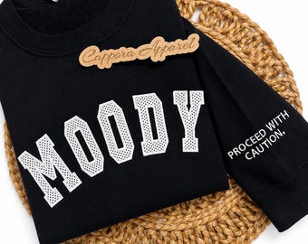 Puede incluir: Una sudadera negra de manga larga con la palabra MOODY en letras grandes blancas de estilo universitario con textura perforada. La manga derecha muestra el texto blanco PROCEED WITH CAUTION. Una etiqueta de marca de madera dice Cappora Apparel.