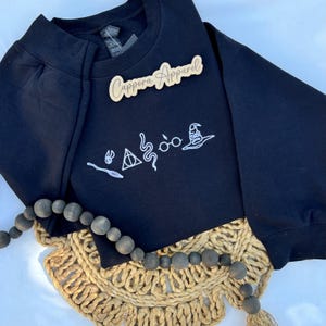 May include: A black long-sleeve sweatshirt featuring white embroidered line art icons including a wand, a snitch, a triangle symbol, a snake, round glasses, and a wizard hat. A wooden sign with the text Cappora Apparel is placed on the fabric.