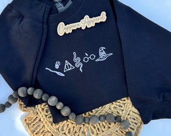 Puede incluir: Una sudadera negra de manga larga con bordados en hilo blanco que representan una varita, una snitch, un símbolo triangular, una serpiente, gafas redondas y un sombrero de mago. Un letrero de madera con el texto Cappora Apparel está sobre la tela.