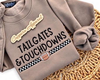 Puede incluir: Una sudadera de cuello redondo color topo con el texto bordado en negro TAILGATES & TOUCHDOWNS. Debajo del texto hay una franja a cuadros blancos y negros con un gráfico de balón de fútbol americano marrón y blanco. Una etiqueta de madera dice Cappara Apparel.