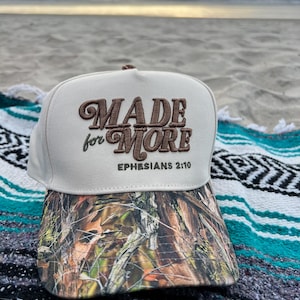 以下が含まれることがあります： ベージュ色の野球帽。茶色で「MADE for MORE」と刺繍されています。帽子のつばは迷彩柄です。「Ephesians 2:10」の文字が下にあります。ビーチの毛布の上に帽子が置かれています。