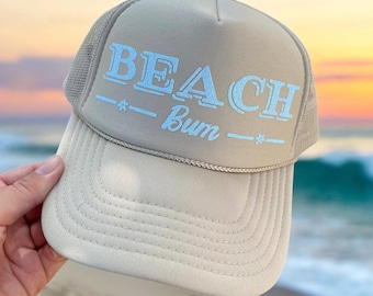 Op de afbeelding: Een beige truckercap met een blauw geborduurd ontwerp met de tekst "BEACH Bum" met sterren aan weerszijden van de tekst.