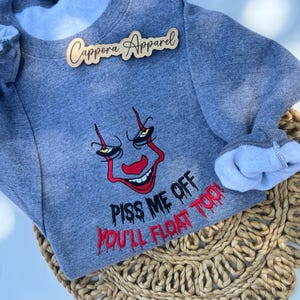 May include: A heather gray crewneck sweatshirt featuring an embroidered graphic of a clown face with yellow eyes and red accents. Below the face, the text reads PISS ME OFF in black and YOU'LL FLOAT TOO! in red. A wooden logo tag reads Cappora Apparel.