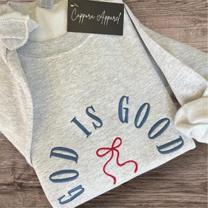 May include: A light gray sweatshirt with the embroidered text "GOD IS GOOD" in navy blue and a red bow. The sweatshirt has a tag that says "Cappora Apparel". The garment is folded on a wooden surface.
