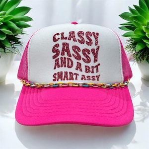 May include: A pink and white trucker hat with a pink glitter design that says "Classy Sassy and a Bit Smart Assy". The hat has a colorful chain trim.