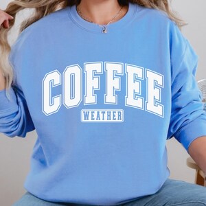 Op de afbeelding: Een lichtblauw sweatshirt met de tekst "COFFEE WEATHER" in wit. De tekst is in een groot, vet lettertype.