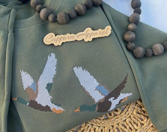 Puede incluir: Una sudadera de color verde oliva con un bordado de punto de cruz de dos patos en vuelo. El diseño incluye tonos marrones, blancos, azules y naranjas. Un letrero de madera con el texto Cappora Apparel se sitúa sobre los patos, y un collar de cuentas de madera oscura se extiende por la parte superior.