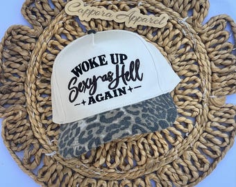 Puede incluir: Una gorra de béisbol color crema con visera plana que presenta un estampado de leopardo en gris y negro. La parte frontal de la gorra muestra un texto bordado en negro que dice WOKE UP SEXY AS HELL AGAIN con pequeñas estrellas a los lados.