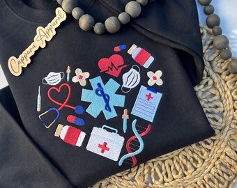 Puede incluir: Una sudadera negra con un bordado en forma de corazón compuesto por iconos médicos, incluyendo un estetoscopio, una estrella de la vida azul, un botiquín de primeros auxilios, una cadena de ADN, tiritas, pastillas, un portapapeles y mascarillas.