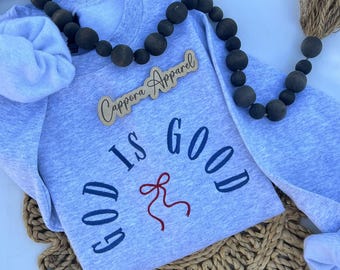 Könnte beinhalten: Ein hellgraues Sweatshirt mit Rundhalsausschnitt und dunkelblau gesticktem Text God Is Good sowie einer kleinen roten Schleifengrafik in der Mitte. Ein Holzschild mit der Aufschrift Cappora Apparel liegt über dem gestickten Design.