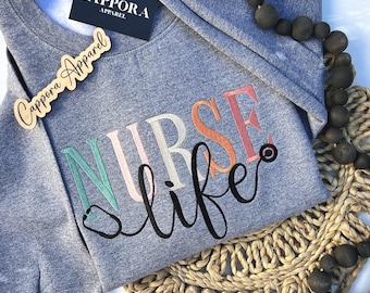 Puede incluir: Una sudadera gris jaspeado de cuello redondo con la palabra NURSE en letras de bloque multicolores y la palabra life en cursiva negra con un gráfico de estetoscopio integrado. Una etiqueta negra muestra CAPPORA APPAREL.