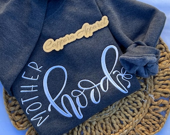 Puede incluir: Una sudadera de manga larga gris carbón oscuro con texto bordado en blanco que dice "MOTHER" verticalmente y "hood" horizontalmente en letra cursiva. Una etiqueta de madera con "Cappora Apparel" está colocada encima.