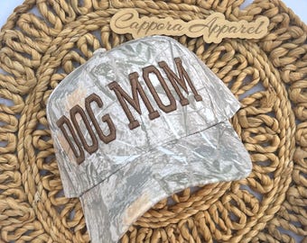 Puede incluir: Una gorra de béisbol con un patrón de camuflaje en tonos grises, blancos y verdes. La parte frontal de la gorra muestra el texto DOG MOM bordado en letras mayúsculas de color marrón. Se ve un letrero de madera con el texto Cappora Apparel.