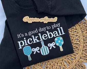 Puede incluir: Una sudadera negra de manga larga con el texto bordado 'it's a good day to play pickleball' en blanco, acompañada de tres palas de pickleball en azul verdoso y verde, y dos lazos blancos. Se incluye una etiqueta de madera 'Cappora Apparel'.
