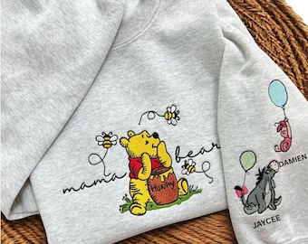 Puede incluir: Una sudadera gris claro de cuello redondo con un bordado de Winnie the Pooh sosteniendo una olla de miel etiquetada como Hunny, rodeado de abejas y el texto mama bear. La manga presenta a Piglet con un globo y a Eeyore bordados con nombres.