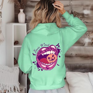 May include: A light green hoodie with a graphic on the back featuring purple lips, a jack-o'-lantern, and the words "Basic Witch." The design includes purple splatters and small bat silhouettes.