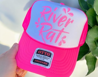 May include: A neon pink trucker hat with a white foam front panel featuring the text River Rat in glittery pink script. The hat includes a black and white sticker on the brim with the text OTTO World Famous Foam Front Trucker OSFM.