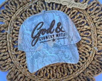 Puede incluir: Una gorra de béisbol con un patrón de camuflaje gris claro y blanco. La parte frontal presenta un texto bordado en marrón que dice God & COUNTRY MUSIC con una pequeña estrella. Arriba, un letrero de madera muestra el texto Cappora Apparel.