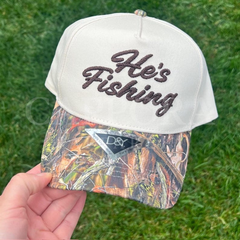 Puede incluir: Gorra de b&eacute;isbol color crema con el texto bordado "He's Fishing" en escritura marr&oacute;n. La visera presenta un patr&oacute;n de camuflaje en tonos marrones, verdes y beige. La gorra tiene un bot&oacute;n en la parte superior y una etiqueta en la visera.
