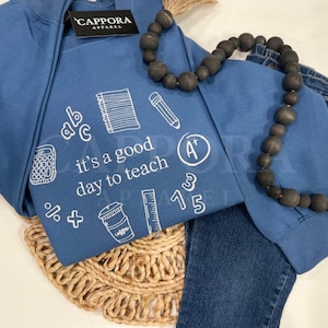 Op de afbeelding: Een blauwe sweatshirt met de tekst "it's a good day to teach" en schoolgerelateerde afbeeldingen. Een zwarte kralenketting en een spijkerbroek staan ook op de foto. De sweatshirt heeft een label met "CAPPORA APPAREL".