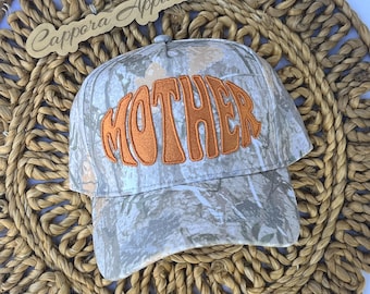 Puede incluir: Una gorra de béisbol con un patrón de camuflaje en tonos grises, blancos y verdes. La parte frontal de la gorra presenta la palabra MOTHER bordada en letras grandes de color naranja. Se observa un letrero de madera con el texto Cappora Apparel.