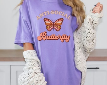 May include: A light purple short-sleeve t-shirt featuring a graphic of a butterfly with orange, pink, and brown wings. The text "ANTI-SOCIAL" is printed in a curved arch above the butterfly, and the word "Butterfly" is printed below in a script font.