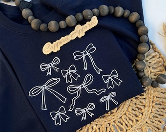 Puede incluir: Una sudadera azul marino de cuello redondo con un gráfico de líneas blancas de varios lazos dispersos por el frente. Una etiqueta de madera con el texto Cappora Apparel se sitúa cerca del cuello, junto a un collar de cuentas de madera oscura.