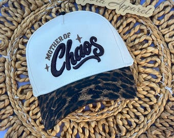 Puede incluir: Una gorra de béisbol color crema con el texto bordado Mother of Chaos en letra cursiva marrón oscuro, acentuado por dos pequeñas estrellas. La gorra tiene una visera curva con un estampado de leopardo marrón y negro y un botón superior.