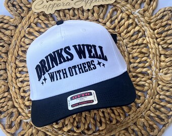 Puede incluir: Una gorra de béisbol blanca con visera negra y un botón negro en la parte superior. El frente presenta un texto bordado en negro que dice DRINKS WELL WITH OTHERS, acompañado de dos pequeñas estrellas de cuatro puntas. Una etiqueta está en la visera.