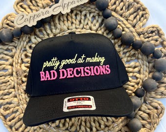 Puede incluir: Una gorra de béisbol negra con el texto bordado en amarillo "pretty good at" sobre el texto en rosa "BAD DECISIONS". La gorra tiene una corona estructurada, una visera plana y una pegatina rectangular en la visera que dice "OTTO CLASSIC FIT".