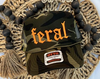 Puede incluir: Una gorra de camionero de camuflaje con un panel frontal de espuma que presenta la palabra feral bordada en letras góticas naranjas. La gorra incluye una parte trasera de malla, una visera curva y una pegatina blanca con el texto OTTO TRUCKER.