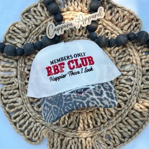 Könnte beinhalten: Eine weiße Baseballkappe mit einem Schirm im Leopardenmuster. Auf der Vorderseite befindet sich ein in Schwarz und Rot gestickter Text: MEMBERS ONLY RBF CLUB Happier Than I Look. Ein kleines D&Y-Markenetikett ist mittig am Schirm befestigt.