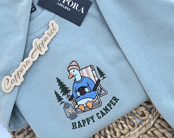 Puede incluir: Una sudadera de cuello redondo color azul claro con un bordado de un ganso con gorro beige y suéter azul, sentado en una silla plegable con una taza, una linterna y pinos. El texto HAPPY CAMPER está bordado en la parte inferior.