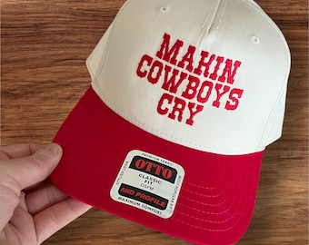 May include: A white and red baseball cap with the text "MAKIN COWBOYS CRY" embroidered in red. The cap has a red brim and a red button on top. The cap is a classic fit and mid profile.