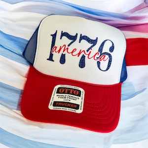 May include: A red, white, and blue trucker hat featuring a white foam front panel with the dark blue glitter text 1776 and the red cursive text america. The hat has a red brim, blue mesh back panels, and a white sticker on the brim with black text.