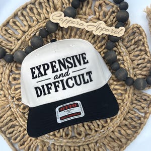 Peut inclure: Une casquette de baseball blanche avec une visière noire portant le texte brodé en noir EXPENSIVE and DIFFICULT. Un autocollant rectangulaire sur la visière affiche le texte OTTO CLASSIC FIT OSFM MID PROFILE MAXIMUM COMFORT. Un panneau en bois indique Cappora Apparel.