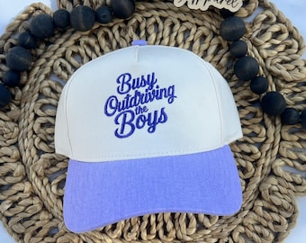 Puede incluir: Una gorra de béisbol de cinco paneles con una corona color blanco hueso y una visera de color morado claro. La parte frontal de la corona presenta un texto bordado en morado oscuro que dice Busy Outdriving the Boys en cursiva.