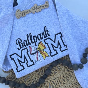 May include: A light grey heathered sweatshirt featuring the black embroidered text Ballpark MOM, with the letter O replaced by a yellow and white baseball-themed bow graphic. A wooden sign with the text Cappora Apparel is placed at the top.