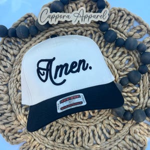 May include: A white baseball cap with a black brim and the word Amen embroidered in black script on the front. A rectangular sticker on the brim reads Otto Classic Fit OSFM. The cap is positioned on a woven tan mat with dark wooden beads.
