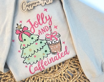 Puede incluir: Una sudadera beige con un diseño bordado de un árbol de Navidad verde con un lazo rosa y una taza de café marrón con un lazo rosa y pajita. El texto dice Jolly AND Caffeinated en rosa. Un letrero de madera muestra Cappora Apparel.