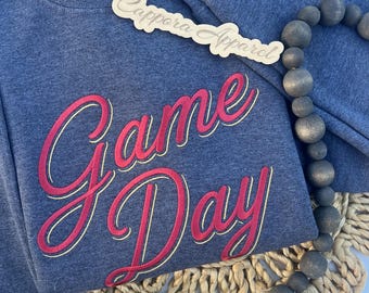 Puede incluir: Una sudadera azul oscuro jaspeado con el texto "Game Day" en rojo con un borde dorado. Un letrero de madera recortado con el texto "Cappora Apparel" se coloca encima. Un collar de cuentas de madera gris oscuro está colocado a un lado.