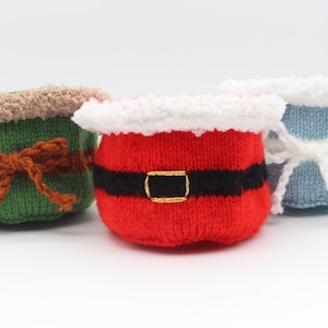 May include: Three knitted Christmas-themed containers. One is green with a brown bow, another is red with a black belt and gold buckle, and the last is blue and white with a white bow. The containers have a fluffy trim.