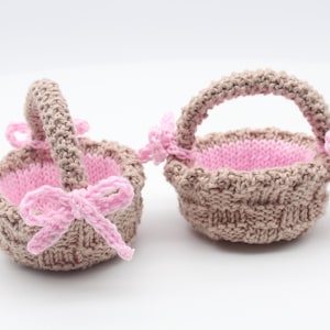 May include: Two small, hand-knitted baskets. The baskets are light brown with pink interiors and pink bows. Each basket has a handle. The baskets are on a white background.