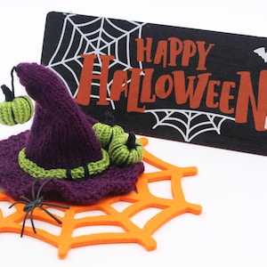 May include: Purple knitted witch hat with green trim and two small green pumpkins. The hat is sitting on an orange spiderweb. A black sign with white spiderwebs and the text "Happy Halloween" is in the background.